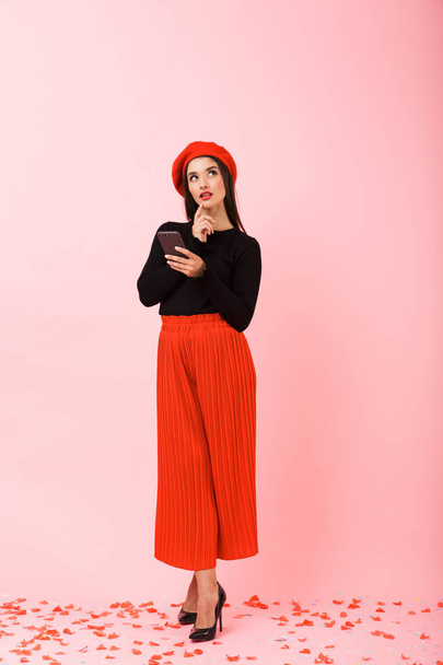 Full length portrait of a pensive beautiful young woman wearing red beret standing isolated over pink background - Fotó, kép