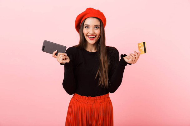 Portrait d'une belle jeune femme heureuse portant béret rouge debout isolé sur fond rose, tenant téléphone portable et carte de crédit en plastique
 - Photo, image