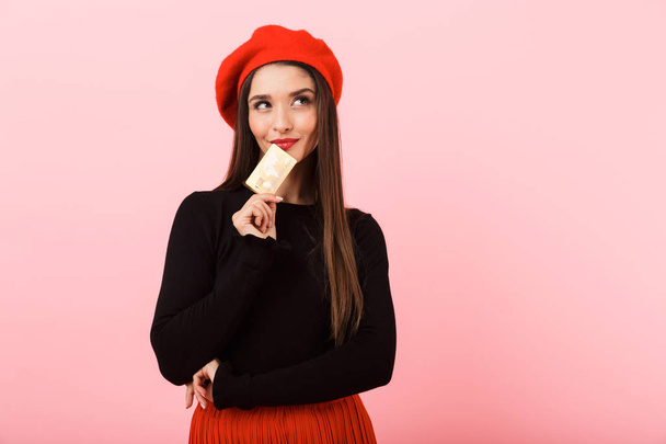 Portrait of a happy beautiful young woman wearing red beret standing isolated over pink background, holding plastic credit card - Valokuva, kuva
