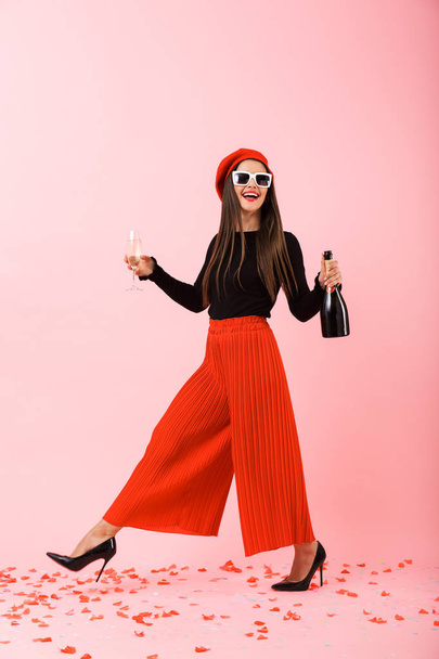 Cheerful young woman wearing sunglasses and red beret celebrating, holding bottle of champagne and a glass - Foto, Imagem