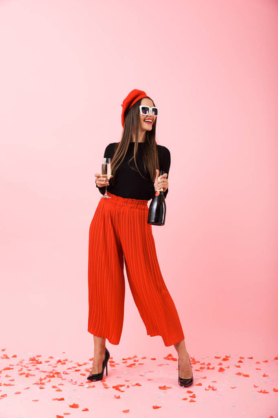 Cheerful young woman wearing sunglasses and red beret celebrating, holding bottle of champagne and a glass - 写真・画像