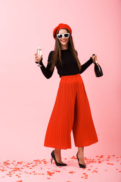 Cheerful young woman wearing sunglasses and red beret celebrating, holding bottle of champagne and a glass - Fotografie, Obrázek