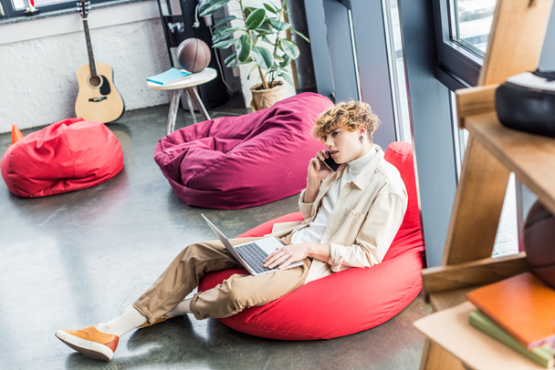 homem de negócios casual bonito sentado na cadeira saco de feijão ao usar laptop e falando no smartphone no escritório loft
 - Foto, Imagem