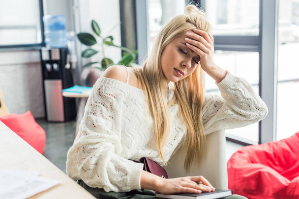 moe casual zakenvrouw zit aan bureau met laptop en met hoofdpijn in loft kantoor - Foto, afbeelding