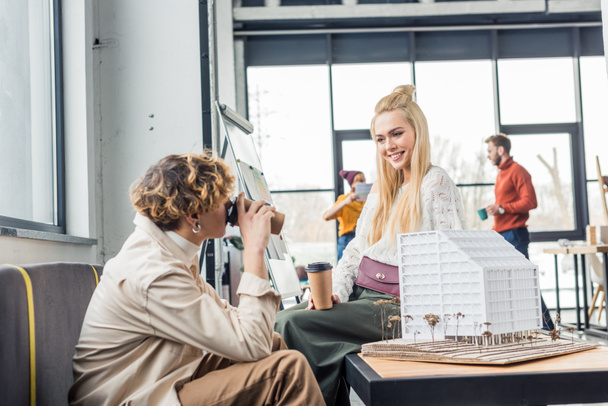 female and male architects with coffee to go looking at each other while working on house model in loft office - Фото, изображение