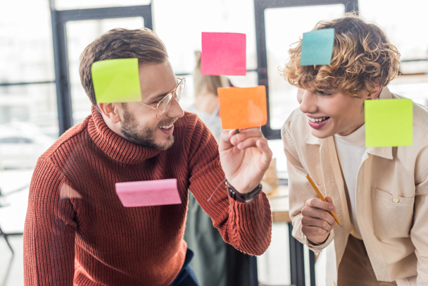 selective focus of handsome casual businessmen putting colorful sticky notes on glass window in office - Foto, Imagem