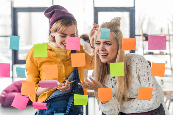 beautiful casual businesswomen laughing near colorful sticky notes on glass window in office - 写真・画像
