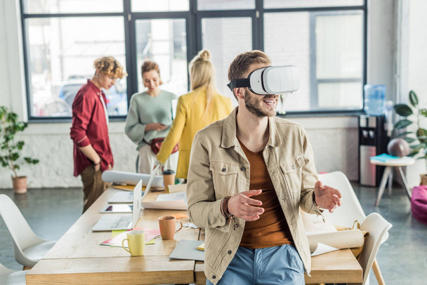 smiling male designer gesturing with hands while having virtual reality experience in loft office with colleagues on background - Foto, afbeelding