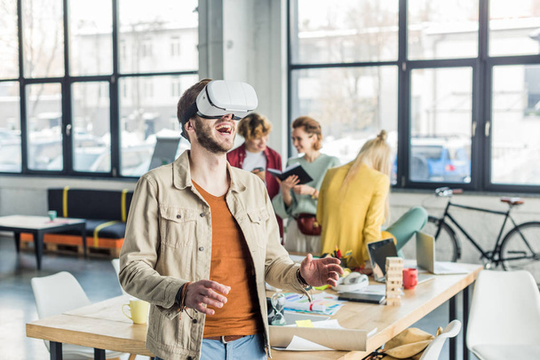 excited male designer gesturing with hands while having virtual reality experience in loft office with colleagues on background - Fotografie, Obrázek