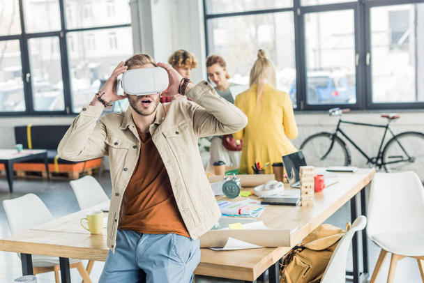 surprised male designer gesturing with hands while having virtual reality experience in loft office with colleagues on background - Foto, afbeelding