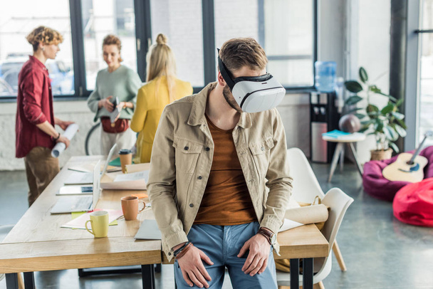 mannelijke ontwerper in vr headset met virtuele werkelijkheid ervaren in loft kantoor met collega's op achtergrond - Foto, afbeelding