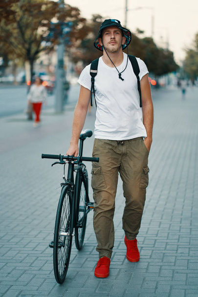 Ritratto all'aperto di un giovane uomo moderno per strada. Un giovane atletico che indossa pantaloni kaki, maglietta bianca, scarpe da ginnastica rosse e zaino alla scoperta della città europea
. - Foto, immagini