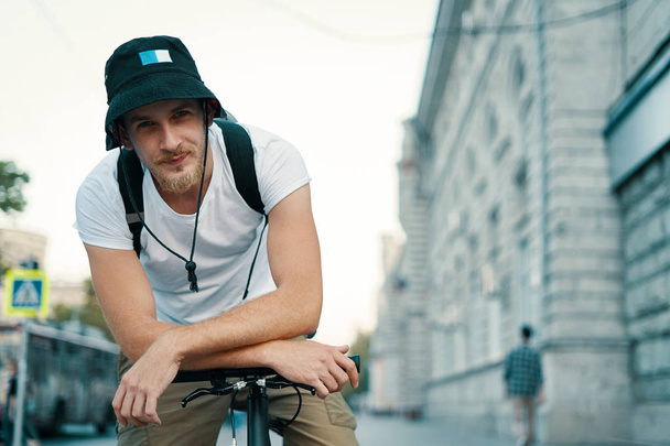 Ένας χαμογελαστός άνθρωπος οδήγηση ενός ποδηλάτου σε μια παλιά Ευρωπαϊκή πόλη που ψάχνει σε εξωτερικούς χώρους κοντά στο σκοπό της κάμερας. Ανδρικό πορτραίτο εκμετάλλευση σταυρωμένα χέρια στη γραμμή λαβή ποδηλάτου. - Φωτογραφία, εικόνα