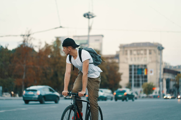Portrait d'un jeune homme en vélo sur la route de la ville, rue avec la ville loin en arrière-plan. Homme sur vélo noir avec chemise blanche, casquette, sac à dos vélo à destination
 - Photo, image
