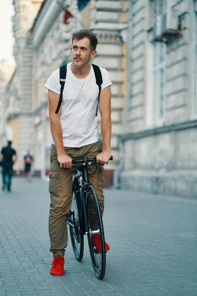 Zewnątrz portret nowoczesne młody człowiek na ulicy, siedząc na rowerze. Młody człowiek lekkoatletycznego na sobie spodnie khaki, biały T-shirt, czerwone trampki i plecak zwiedzanie europejskiego miasta. - Zdjęcie, obraz