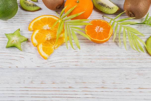 Green palm leaves and fresh tropical slices of fruits on white wooden table. Juicy oranges, tangerines, limes, kiwis, carambolas. Summer fruit background. Flat lay, top view, copy space - Foto, Imagen