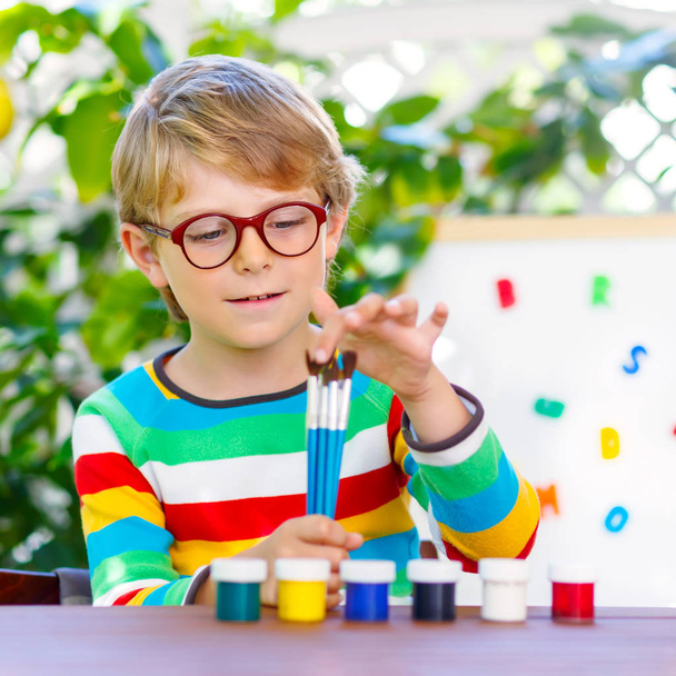 Funny adorable little kid boy with glasses holding watercolors and brushes. Happy child and student is back to school. Education, school, learning concept. School, preschool nursery equipment. - Photo, Image