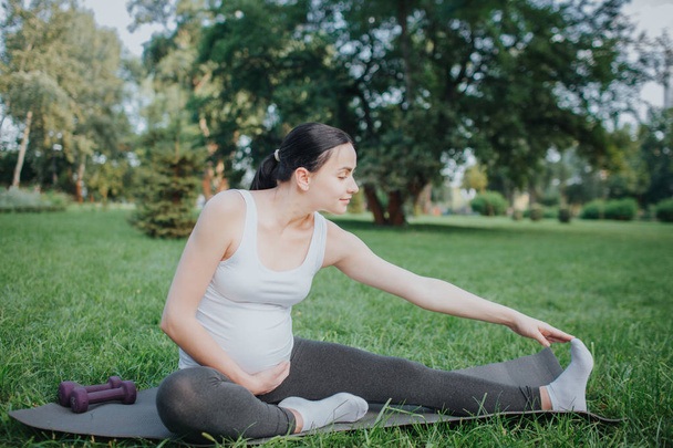 Nice young pregnant woman stretch outside in park. She reaches feet with one hand. Model hold another one on belly. She looks calm and peaceful. - Фото, изображение