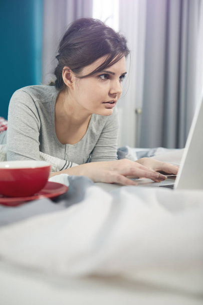 Young woman working with laptop lying on the bed in the bright and cozy bedroom at home - Foto, Bild