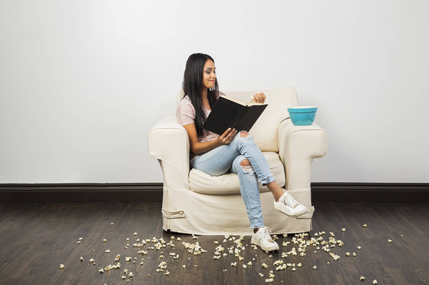молодая женщина сидит на диване, читает книгу и ест попкорн, оставляя беспорядок - Фото, изображение