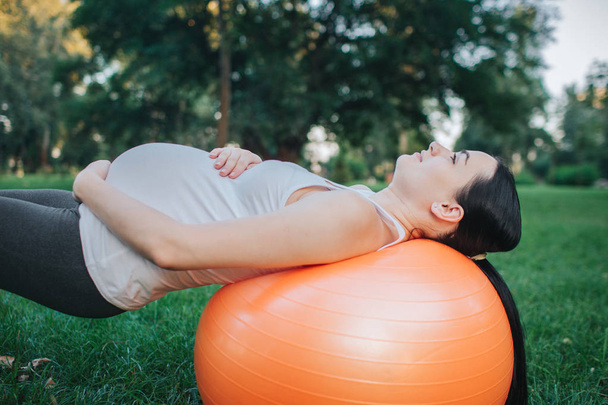 Picture of young pregnant woman lying on orange fitness ball in park. She keep handsarounf belly. Model looks concentrated. - Foto, Imagem