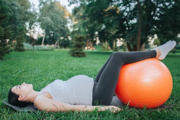 Concentrated young pregnant woman lying on yoga mate in park and look up. She hold legs on orange fintess ball. Model stretch back. - Valokuva, kuva