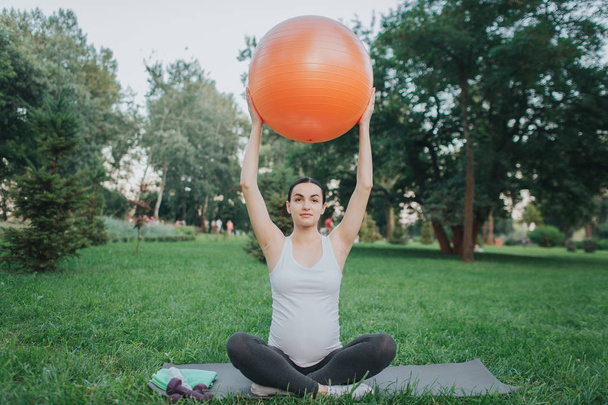 Concentrated young woman sit in lotus pose on yoga mate in park. She hold orange fitness ball up with hands. - Foto, Imagen