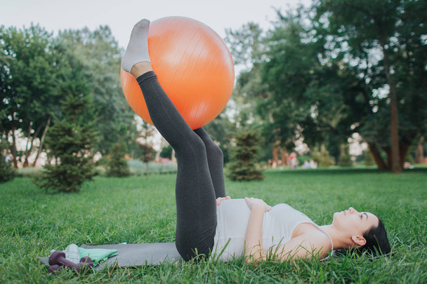Jonge zwangere vrouw exerising geconcentreerd op yoga stuurman in park. Ze houden de grote oranje fitness bal tussen de benen. Model houden handen op buik. - Foto, afbeelding