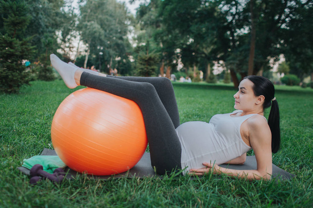Young pregnant woman suffer from pain in hips area. She lying on yoga mate in park. Model hold hands on hips and legs on orange ball. - Foto, afbeelding