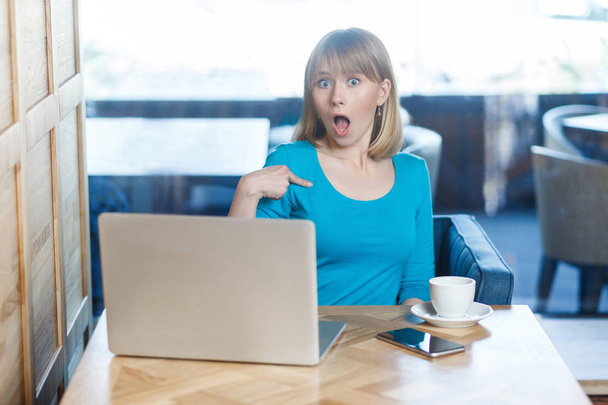 surprised woman in blue blouse pointing finger to herself with big eyes and open mouth while sitting with laptop in cafe, concept of new proposal for work   - Foto, Bild