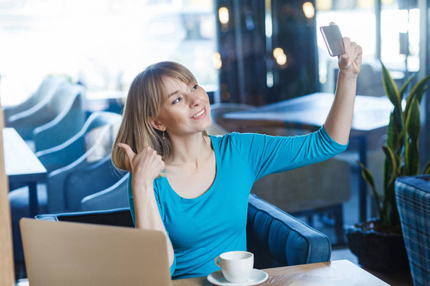 satisfied funny young blonde woman in blue blouse holding smartphone and making selfie while showing thumb up  - Foto, imagen