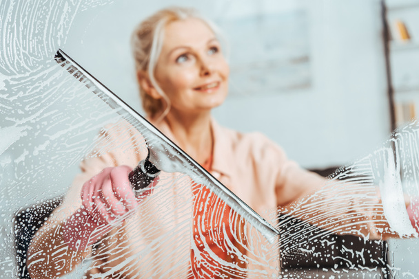 Χαμογελώντας ανώτερων γυναίκα καθαρίζοντας παράθυρο με γυαλί υαλοκαθαριστήρα - Φωτογραφία, εικόνα
