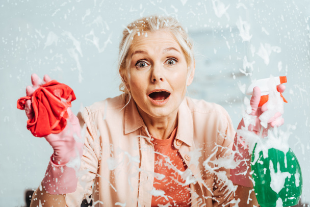 Shocked senior woman in rubber gloves cleaning window - Фото, зображення