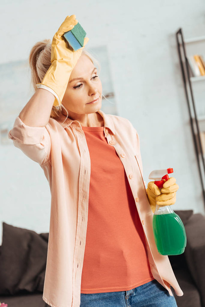 Exhausted senior woman in yellow gloves holding cleaning spray and sponge - Фото, изображение