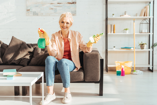 Счастливая женщина в джинсах, сидящая на диване с чистящими средствами после работы по дому
 - Фото, изображение