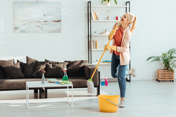 Naurava vanhempi nainen poseeraa yhdellä jalalla puhdistaessaan lattiaa mopilla
 - Valokuva, kuva