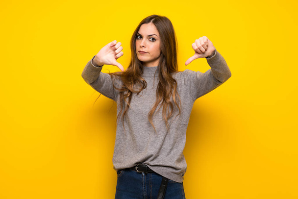 Jovem mulher sobre a parede amarela mostrando polegar para baixo com ambas as mãos
 - Foto, Imagem