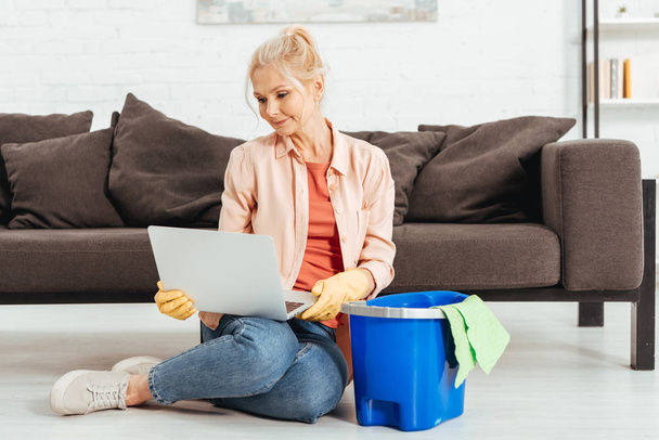 Curious senior woman in rubber gloves posing with laptop while cleaning house - Photo, Image