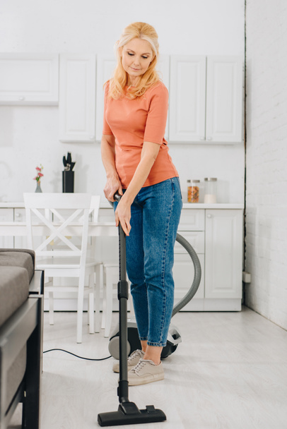 Senior woman in jeans cleaning floor with vacuum cleaner - Fotó, kép