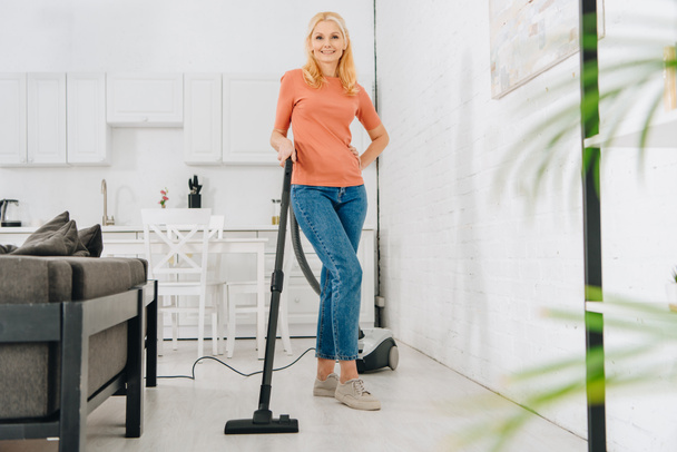 Happy senior woman in jeans cleaning house with vacuum cleaner - Фото, зображення