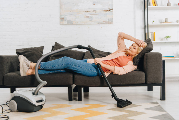 Exhausted woman with vacuum cleaner lying on sofa and touching head - Foto, imagen
