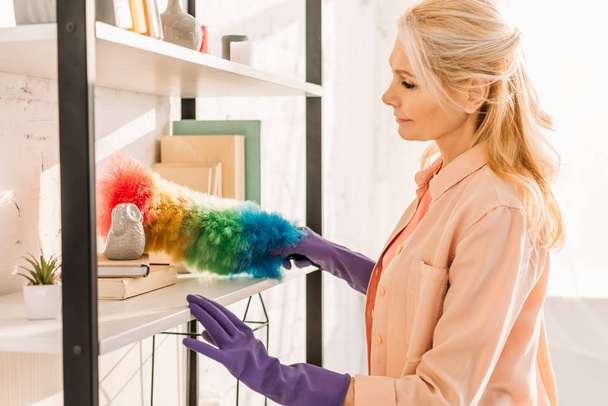 Senior woman in rubber gloves cleaning shelves with duster - Foto, Bild