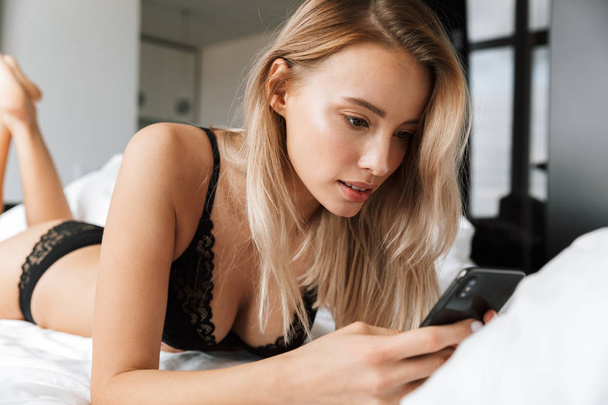 Beautiful smiling young woman wearing lingerie laying in bed, using mobile phone - Φωτογραφία, εικόνα