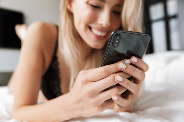 Kép a gyönyörű nő 20s viselése luxus fehérnemű nevetve, miközben feküdt az ágyban, a fehér vászon, hangulatos lakás és használ smartphone - Fotó, kép