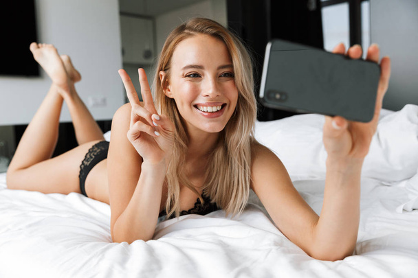 Beautiful smiling young woman wearing lingerie laying in bed, taking a selfie - Fotografie, Obrázek