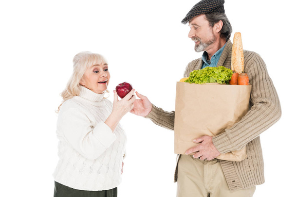 senior man showing apple to cheerful wife while holding paper bag with groceries isolated on white - Фото, зображення