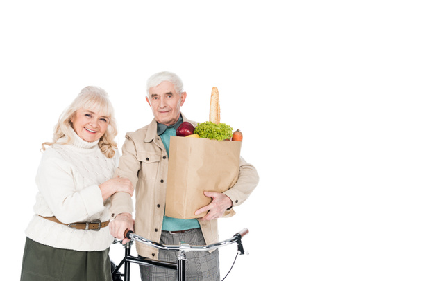 alegre esposa de pie con el marido retirado sosteniendo bicicleta y bolsa de papel aislado en blanco
 - Foto, Imagen