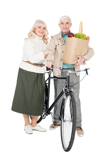 feliz esposa de pie con el marido jubilado sosteniendo bicicleta y bolsa de papel aislado en blanco
 - Foto, Imagen