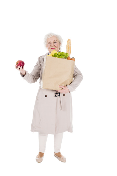 happy woman with grey hair holding apple and paper bag with groceries isolated on white - Foto, Imagem