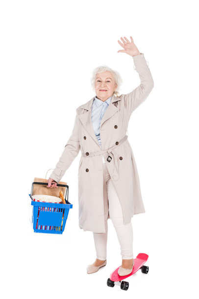 happy senior woman holding shopping basket and waving hand while riding penny board isolated on white - Photo, Image
